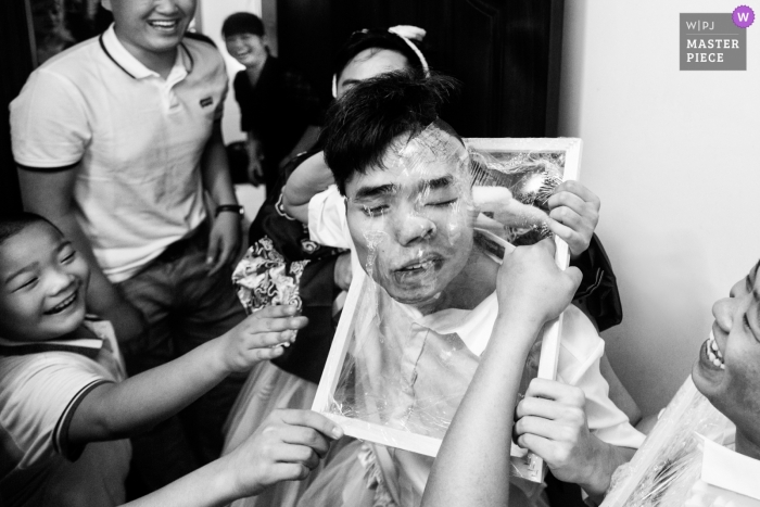 Photo by: <a href="https://www.wpja.com/contests/2019-v04/best-wedding-photographers/2469721">Joy Qin - Longyan </a> Black and white wedding photojournalism with guests molding a plastic frame around young man's face in Fujian, China.