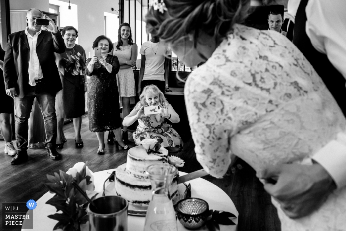 Photo by: <a href="https://www.wpja.com/contests/2019-v06/best-wedding-photographers/2485901">Lubow Polyanska - Prague </a> A woman takes a picture with her phone as the bride and groom cut their cake in this black and white photo created by a Prague documentary wedding photographer.
