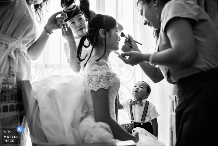 Bridesmaids e un ragazzino guardano mentre la sposa si fa truccare in questa foto in bianco e nero di un fotografo di matrimoni di Pechino, in Cina.