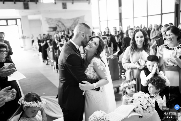 Photo by: <a href="https://www.wpja.com/contests/2019-v07/best-wedding-photographers/2500866">Danilo Muratore - Rosarno Reggio Calabria</a> Guests watch as the groom kisses the bride on the cheek in Gioia Tauro in this black and white award-winning photo by a Calabria wedding photographer.