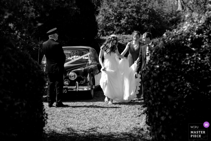 Foto von: <a href="https://www.wpja.com/contests/2019-v07/best-wedding-photographers/2501511"> Catherine Hill - Canterbury Kent </a> Hayne House, Kent, UK Hochzeitsfotografie | Die Braut kommt unter windigen Bedingungen an, als ihr Vater und ihre Brautjungfer mit ihrem Kleid helfen