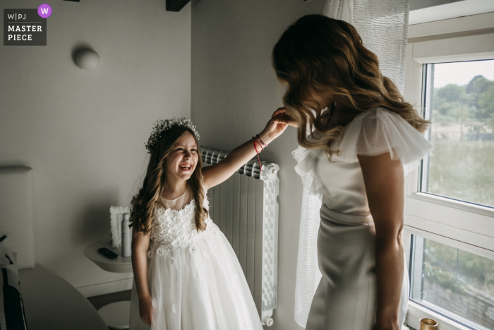 Foto von: <a href="https://www.wpja.com/contests/2019-v07/best-wedding-photographers/2505901"> Giuseppe De Angelis - Norma Latina </a> Die Braut hebt den Arm des Blumenmädchens, um sich vor ihrer Hochzeit im Le Chiacchiere Artena ein Bild von ihrem Kleid zu machen.