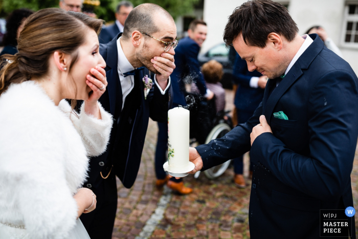 Photo: <a href="https://www.wpja.com/contests/2019-v08/best-wedding-photographers/2509881"> Michael Geyer - Markdorf </a> Les mariés se couvrent la bouche avec surprise en réalisant qu'ils ont accidentellement soufflé de la cire chaude sur le costume de leur invité en soufflant leur bougie de mariage à Fischbach sur cette photo prise par un photographe de mariage de Markdorf.