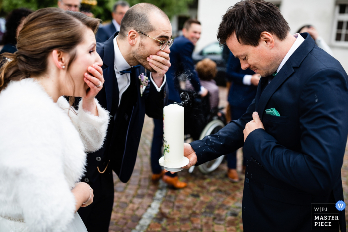 The bride and groom cover their mouths in surprise realizing they accidentally blew hot wax on their guest's suit when blowing out their wedding candle in Fischbach in this photo by a Markdorf wedding photographer. 