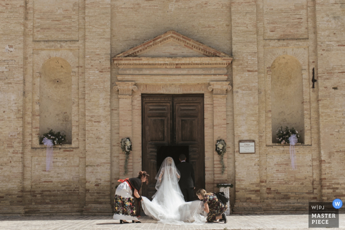 Dos mujeres ayudan a la novia a arreglar su hermoso vestido mientras se prepara para ingresar a la ceremonia en Chiesa Santa Croce Mogliano en esta imagen creada por una fotógrafa de bodas de Macerata, Marche.