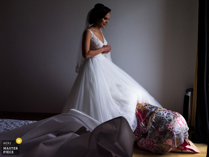 Photo by: <a href="https://www.wpja.com/contests/2019-v08/best-wedding-photographers/2518936">Joshua Dhondt - Antwerpen</a> A woman is partially covered by the bride's dress as she helps the bride get ready at Sint-Michielskerk in this photo by an Antwerpen, Flanders wedding photographer.