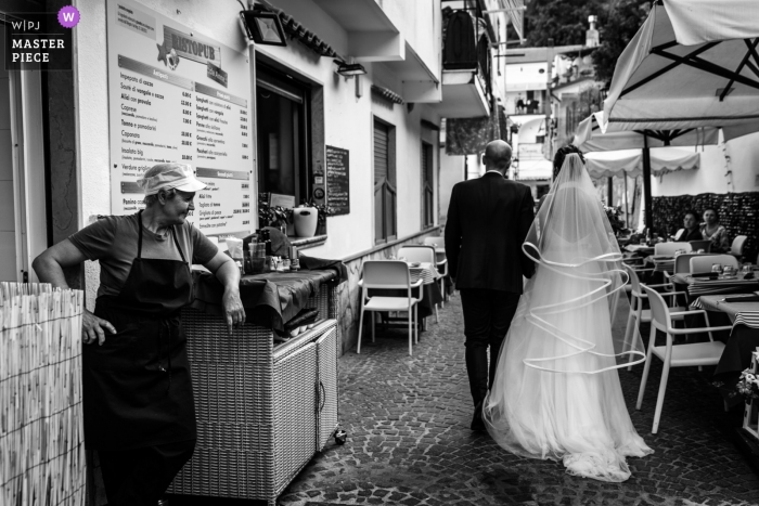 Photo by: <a href="https://www.wpja.com/contests/2019-v09/best-wedding-photographers/2525091">Federica Ariemma - Napoli Napoli</a> A wedding bridal walk through Erchie as a restaurant vendor looks on.