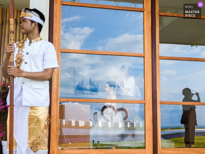 Zdjęcie: <a href="https://www.wpja.com/contests/2019-v09/best-wedding-photographers/2530271"> Leon Wong - Hangzhou </a> Wyspa Fantasy, Bali, Indonezja Zdjęcia z miejsca ślubu - Panna młoda w szatni miejsca ceremonii, ekran naprawił gości i obszar ceremonii w odbiciu okna, a także lokalny personel.