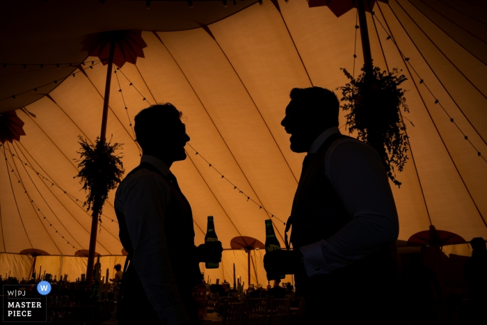 Photo by: <a href="https://www.wpja.com/contests/2019-v09/best-wedding-photographers/2537531">Tom Beynon - Cambridge Cambridgeshire</a> Park Farm, Daventry Tent Wedding Photography - Two guests stand in the marquee to escape the heat outside