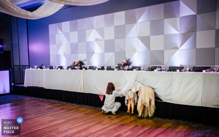 Photo by: <a href="https://www.wpja.com/contests/2019-v09/best-wedding-photographers/2539186">Andrea Bibeault - Omaha Nebraska</a> Photo showing little girls trying to hide under the head table at Scott Conference Center