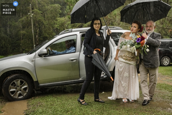 Photo by: <a href="https://www.wpja.com/contests/2019-v09/best-wedding-photographers/2541496">Clara Sampaio - Rio de Janeiro Rio de Janeiro</a> Trilhas do Araçari, Nova Friburgo, Rio de Janeiro - Bride getting out of car in the rain under umbrellas.