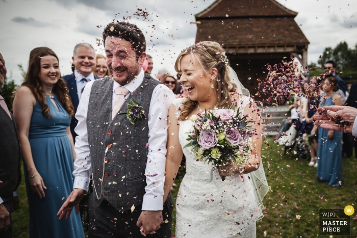 Na een mooie en emotionele huwelijksceremonie in Redcoats Farmhouse loopt het echtpaar het gangpad onder een storm van confetti, inclusief een nieuwe lading die vanaf de rechterkant van de foto binnenkomt. De reacties op hun gezichten zijn geweldig.