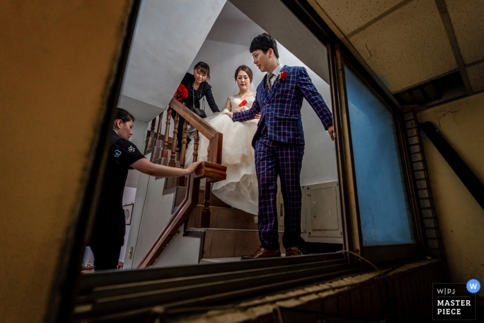 Trouwdagfoto van de bruid die de arm van de bruidegom vasthoudt om de trap af te lopen in het oude appartement van hun ouders in Taiwan.