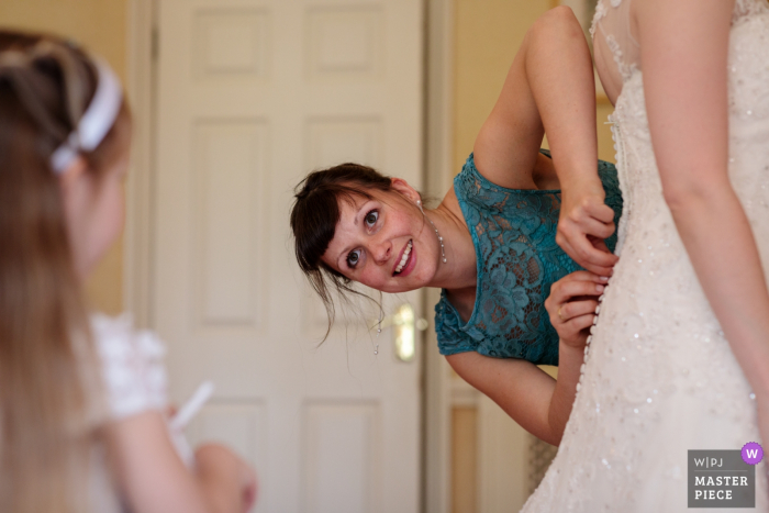 Photo by: <a href="https://www.wpja.com/contests/2019-v10/best-wedding-photographers/2543772">Catherine Hill - Canterbury Kent</a> The bridesmaid does up the buttons to the brides dress whilst her daughter looks on - Wedding Photography from Broome Park, Barham, Kent, UK