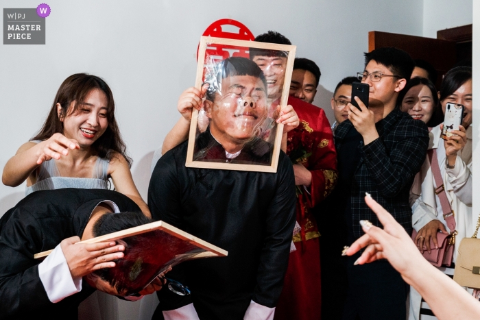 Foto: <a href="https://www.wpja.com/contests/2019-v11/best-wedding-photographers/2545720"> Vincent Mu - De Zhou </a> Die Spielverbindung der Rezeption des Bräutigams. Bräutigam lässt sein Gesicht für eine Hochzeit in China durch einen mit Zellophan umwickelten Rahmen schlagen.