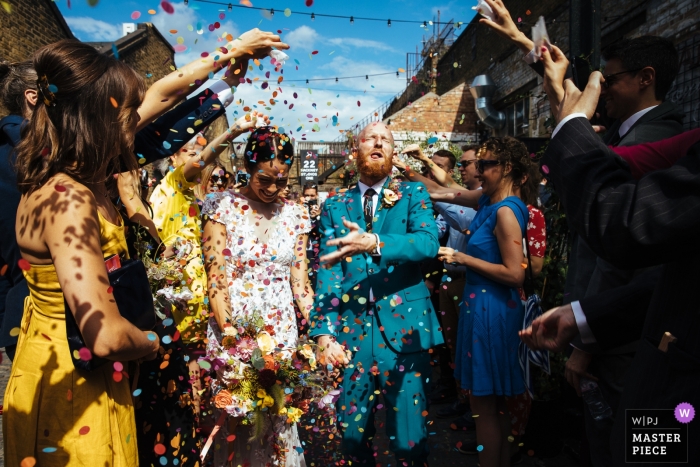 Photo by: <a href="https://www.wpja.com/contests/2019-v12/best-wedding-photographers/2548079">Lyndsey Goddard - London London</a> England wedding reportage photography - Bride and groom exit the ceremony as their guests congratulate them and the groom gets a mouthful of confetti