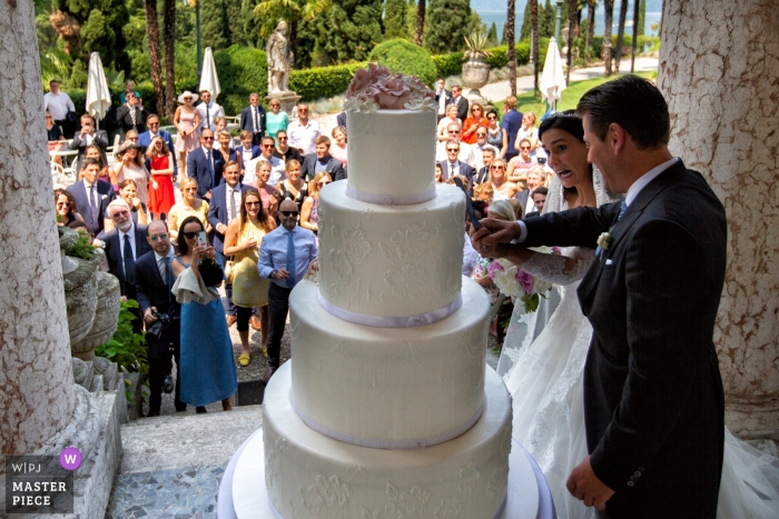 Foto: <a href="https://www.wpja.com/contests/2019-v12/best-wedding-photographers/2548450"> Daniele Borghello - Padua </a> Hochzeitsfotografie von draußen im Villa Cortine Palace Hotel - Sirmione - Gardasee - Italien - Bild des lustigen Kuchenausschnitts mit den Gästen im Hintergrund