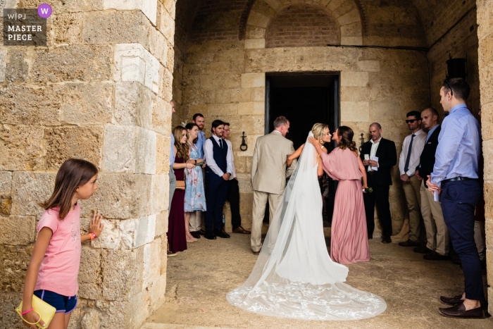 Photo by: <a href="https://www.wpja.com/contests/2019-v12/best-wedding-photographers/2548566">Annie Kheffache - Dun Laoghaire Dublin</a> San Galgano, Tuscany, Italy Wedding Reportage Photographer - Image of a Tourist child peeping at bride outside church preparing to walk in, bridesmaid air kiss.
