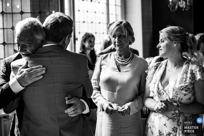 Photo by: <a href="https://www.wpja.com/contests/2019-v13/best-wedding-photographers/2550259">David Pommier - Lyon </a> Lyon wedding ceremony image of the groom receiving a hug from his father as women look on with emotion.