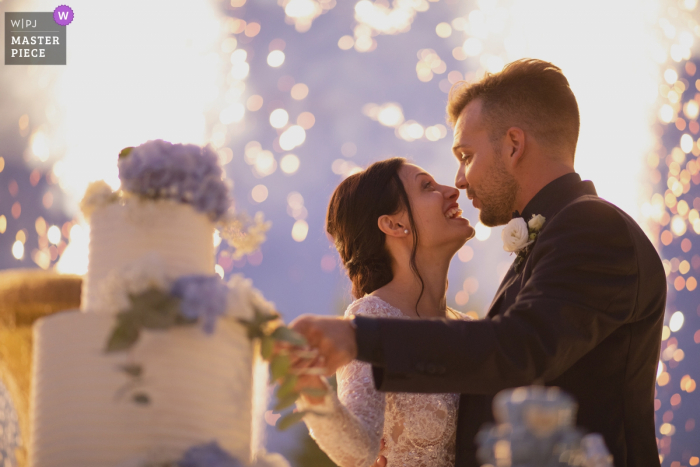 Photo by: <a href="https://www.wpja.com/contests/2019-v14/best-wedding-photographers/2552243">Sabrina Vinci - Civitanova Marche Macerata</a> Villa Castellani wedding photo of the bride and groom cutting the cake with fireworks with in the background.