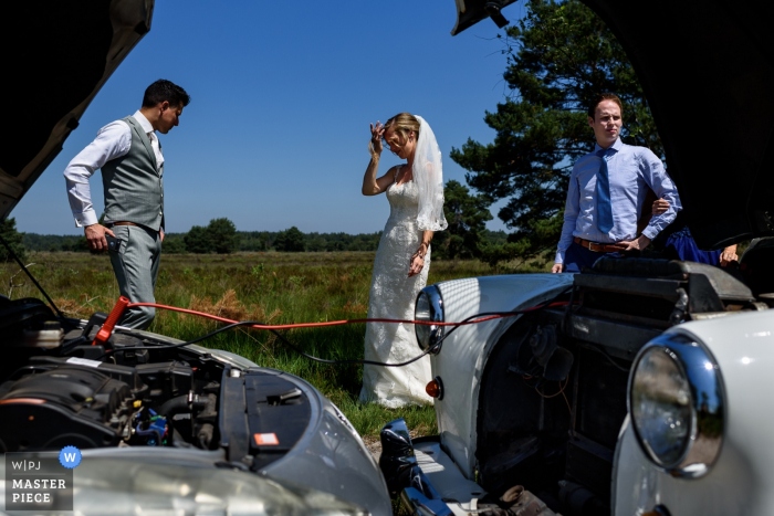 Foto door: <a href="https://www.wpja.com/contests/2019-v14/best-wedding-photographers/2552321"> Elke Teurlings - Geldrop Noord Brabant </a> NL Wedding Fotojournalistiek - De autobatterij van het Wedding Vehicle is leeg. Het wordt opgeladen terwijl de bruidegom net een telefoontje heeft gehad met de ceremonie. De bruid staat met haar hand in haar haar.