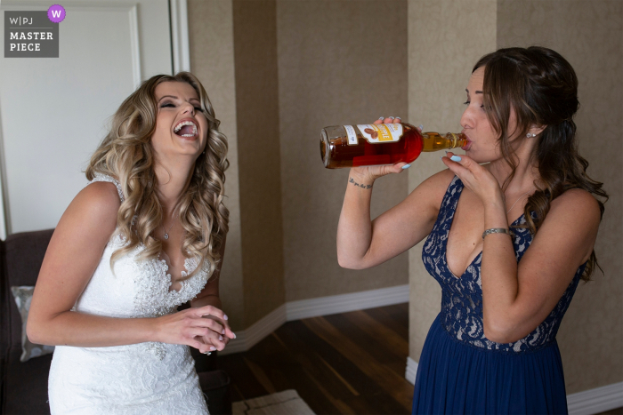 Bruidsmeisje neemt een slok sterke drank terwijl de bruid lacht in een hotel Bruidsmeisje neemt een slokje drank terwijl bruid lacht in een hotel voorafgaand aan de bruiloft in Alberta, Canada.