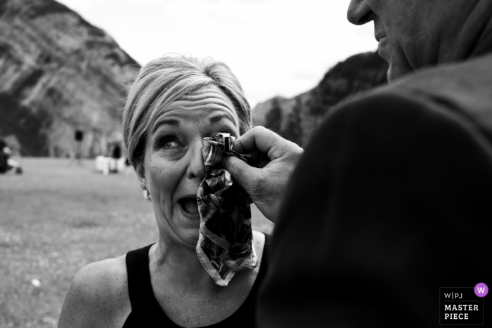 Zwart-witte huwelijksfotografie bij het reservoir van de tunnelberg, Banff, AB, Canada. Vader van de bruid die zijn huilende vrouw (moeder van de bruid) helpt om de tranen te drogen na de emotionele ceremonie.