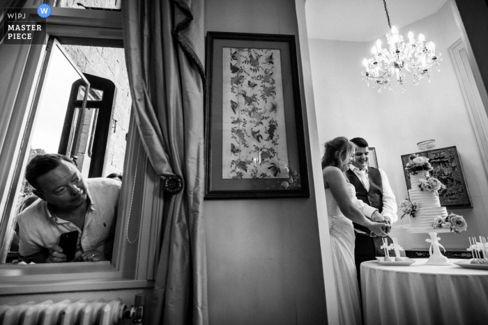 a mirror reflecting the bride and groom cutting the cake, while a man peeks through a window Wadhurst Castle, UK Wedding Venue: photo of a mirror reflecting the bride and groom cutting the cake, while a man peeks through a window.