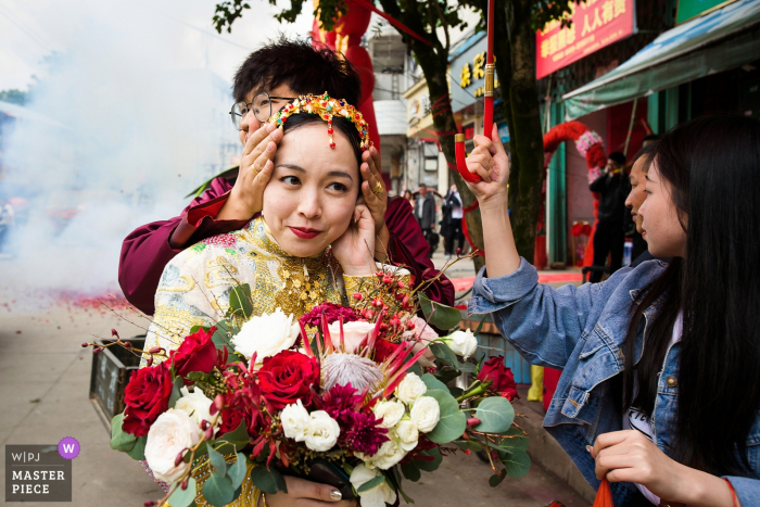 Photo by: <a href="https://www.wpja.com/contests/2019-v15/best-wedding-photographers/2556933">Xiang Liu - Nanping </a> Fujian, China wedding day photo of the bride's ears being covered as firecrackers explode and smoke fills the air.