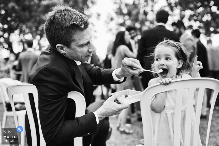 Photo by: <a href="https://www.wpja.com/contests/2019-v15/best-wedding-photographers/2557021">Franck Boutonnet - Lyon </a> During the cocktail session at a wedding a dad is feeding her daughter at the Domaine de Sarson in Grignan, France