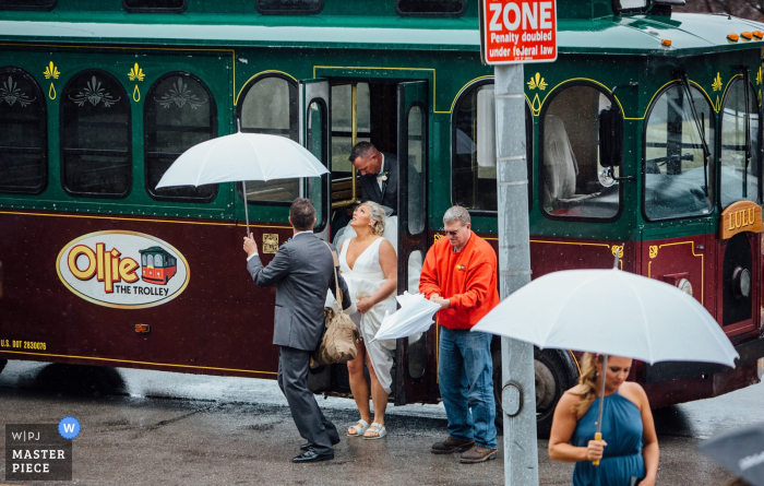 Foto von: <a href="https://www.wpja.com/contests/2019-v16/best-wedding-photographers/2561704"> Andrea Bibeault - Omaha Nebraska </a> Nebraska-Zeremonie-Fotografie - die Braut, die von der Laufkatze oben betrachtet, regnet - Ollie The Trolley aussteigt