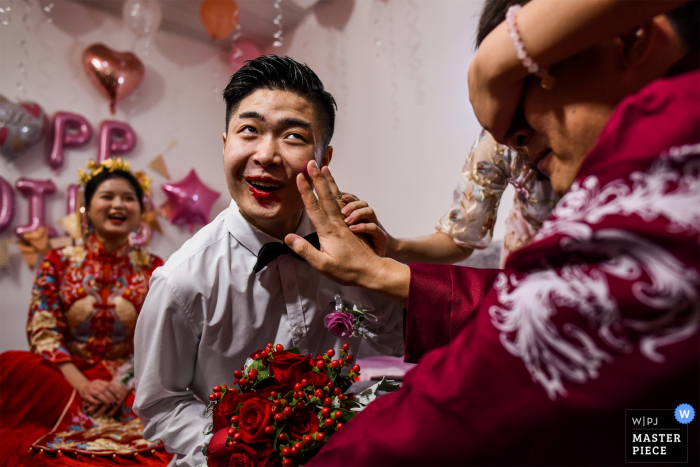 Photo by: <a href="https://www.wpja.com/contests/2019-v17/best-wedding-photographers/2564413">Cheng Guo - ChengDu </a> Chengdu Sichuan wedding photos | Sitting on the bed is the brother of the bridegroom. The sister of the bride covers the eyes of the bridegroom. The bridegroom is making lipstick for his brother. At this moment, his brother's expression is very interestin