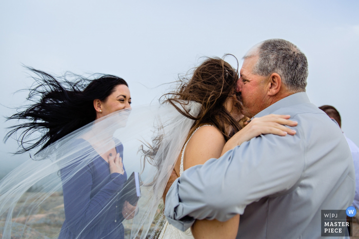 Photo by: <a href="https://www.wpja.com/contests/2019-v17/best-wedding-photographers/2565122">Kate Crabtree - Bangor Maine</a> Cadillac Mountain wedding photos - A father kisses the bride on a windy mountain in Maine