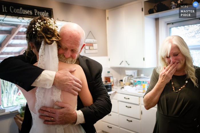 Photo by: <a href="https://www.wpja.com/contests/2019-v19/best-wedding-photographers/2568515">Kent Meireis - Kalispell Montana</a> Brides family farm wedding ceremony and reception site near Kalispell Montana: A bride shares an emotional moment with her parents in the kitchen of the house where they all live before her wedding.