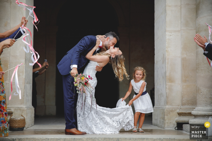 Imagem da cerimônia de casamento em Paris da noiva e do noivo se beijando do lado de fora da igreja após a cerimônia, enquanto uma jovem segura o vestido da noiva com força para que ela desça as escadas.