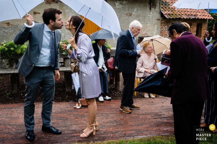 Flanders Kasteel van Brasschaat wedding image contains:  A Quick kiss by guests under umbrellas in the rain.