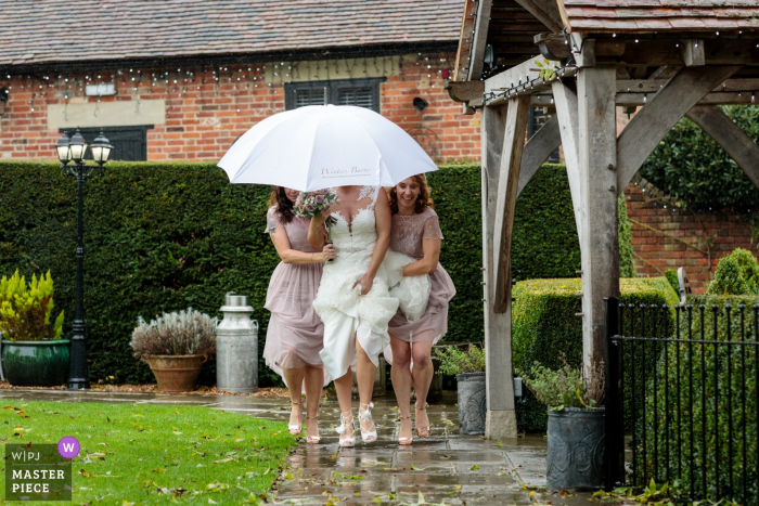 Immagine del reportage di nozze da Winters Barns, Canterbury, Kent, Regno Unito - La sposa è scortata dalle sue damigelle mentre si fa strada per sposarsi e schiva la pioggia