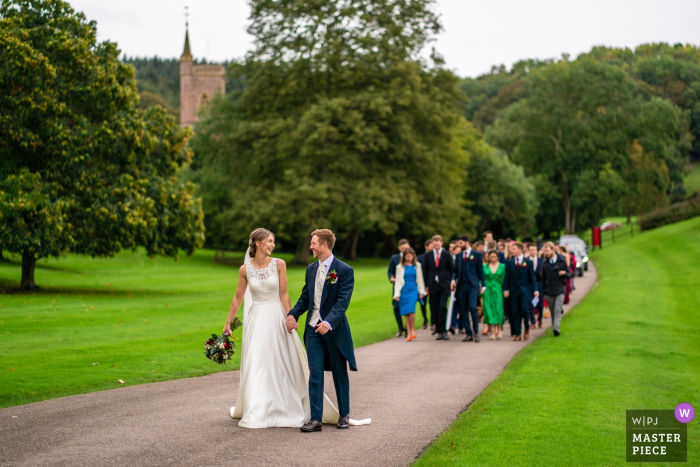 L'immagine del matrimonio di St Audries Park, Somerset, Regno Unito contiene: Sposa e sposo che camminano dalla loro chiesa, sullo sfondo, verso la loro sede, con tutti gli ospiti che seguono