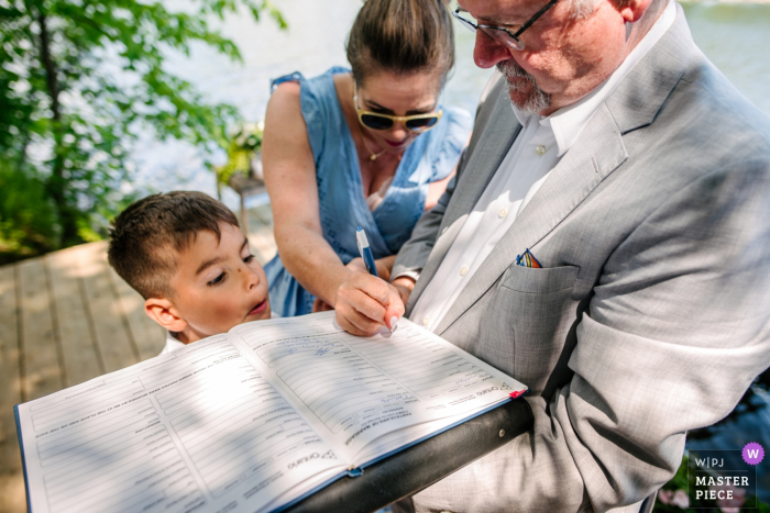 Photo by: <a href="https://www.wpja.com/contests/2019-v19/best-wedding-photographers/2571671">Viara Mileva - Bath Ontario</a> Ontario photography of ceremony at outdoor cottage venue - The boy looks as witnesses sign the papers