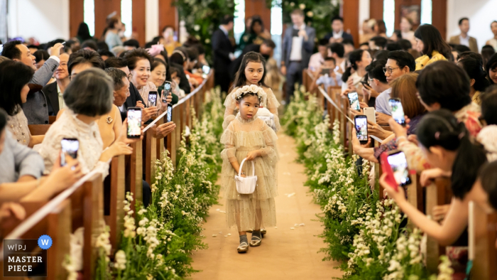 Flowergirls przechadzają się po ołtarzu podczas ceremonii ślubnej w kościele Wattana, podczas gdy wielu gości robi sobie zdjęcia telefonami.