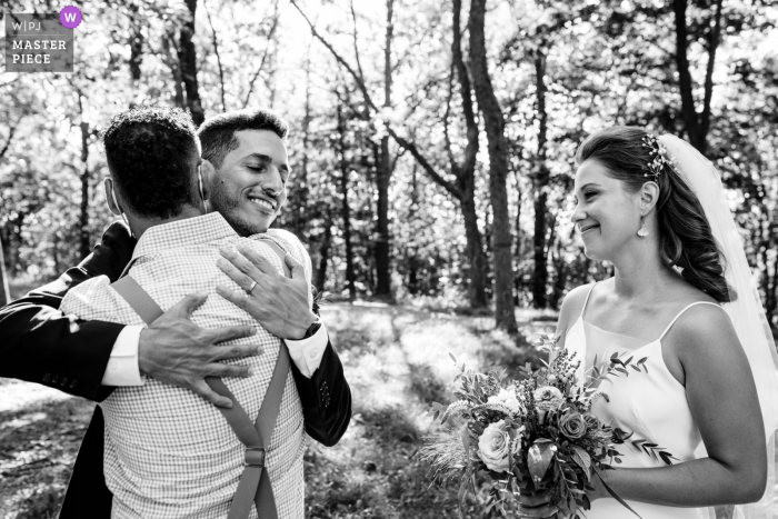 Outdoor wedding photography from Mont Royal Park, Montreal of the Groom being hugged by cousin as bride watches during a micro wedding Outdoor wedding photography from Mont Royal Park, Montreal of the Groom being hugged by cousin as bride watches during a micro wedding