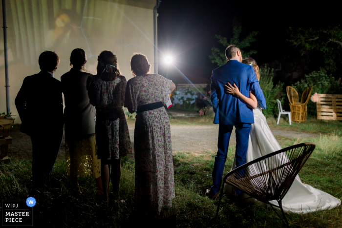 Fotografía de boda en Francia de Vendée con una toma nocturna de la pareja de novios besándose mientras se reproduce una presentación de diapositivas en una pared exterior del lugar