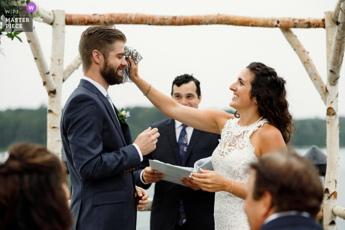 Beach wedding photography from Martha's Vineyard MA at a private house as the Brides wipes the groom's head from rain drops during the ceremony 