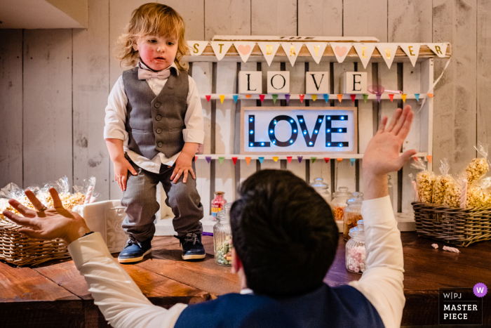 IE fotografia di reportage di matrimonio dai fienili di Segrave, Louth, Irlanda di un papà che cerca di convincere il paggetto bambino a scendere dal carrello delle caramelle