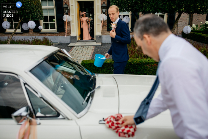 trouwfoto van een huis in Rhenen, Nederland van de bruid die wacht terwijl haar vader en haar bruidegom de auto schoonmaken