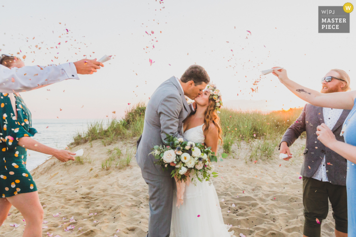 Fotografia de casamento na praia ao ar livre em Clark's Cove, Ilha de Nantucket MA de convidados tomando banho nos recém-casados ​​em confetes de flores secas feitos pela noiva com rosas de sua praia favorita