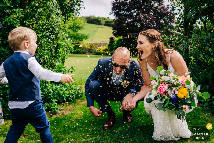 fotografia de casamento da Inglaterra durante a recepção no celeiro Pangdean da noiva e do noivo se agacham rindo enquanto o pajem joga confetes