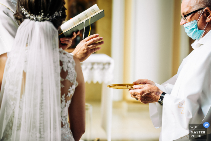 huwelijksfotografie van de Parochie van de Heilige Maagd Maria in Kalisz, Polen van de gemaskerde predikant die de priesterringen op een dienblad overhandigt.