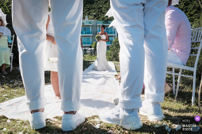 Bruid die door het gangpad loopt bij een particulier opgezette bruiloft voor haar huis. Geschoten door de benen van de bruidegom en het paar in Sai Kung, Hong Kong (Outside the Bride & Groom house, waar ze zich oprichtten voor een privébruiloft vanwege de COVID-19-beperking