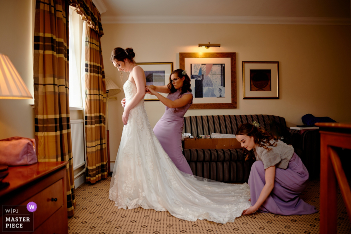 Foto de casamento em Hampshire de uma noiva da Inglaterra se vestindo em seu quarto de hotel com alguma ajuda